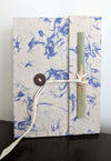 A handmade Lokta paper notebook with blue fiber detailing, secured with a string-and-bamboo tie, standing upright on a dark surface.