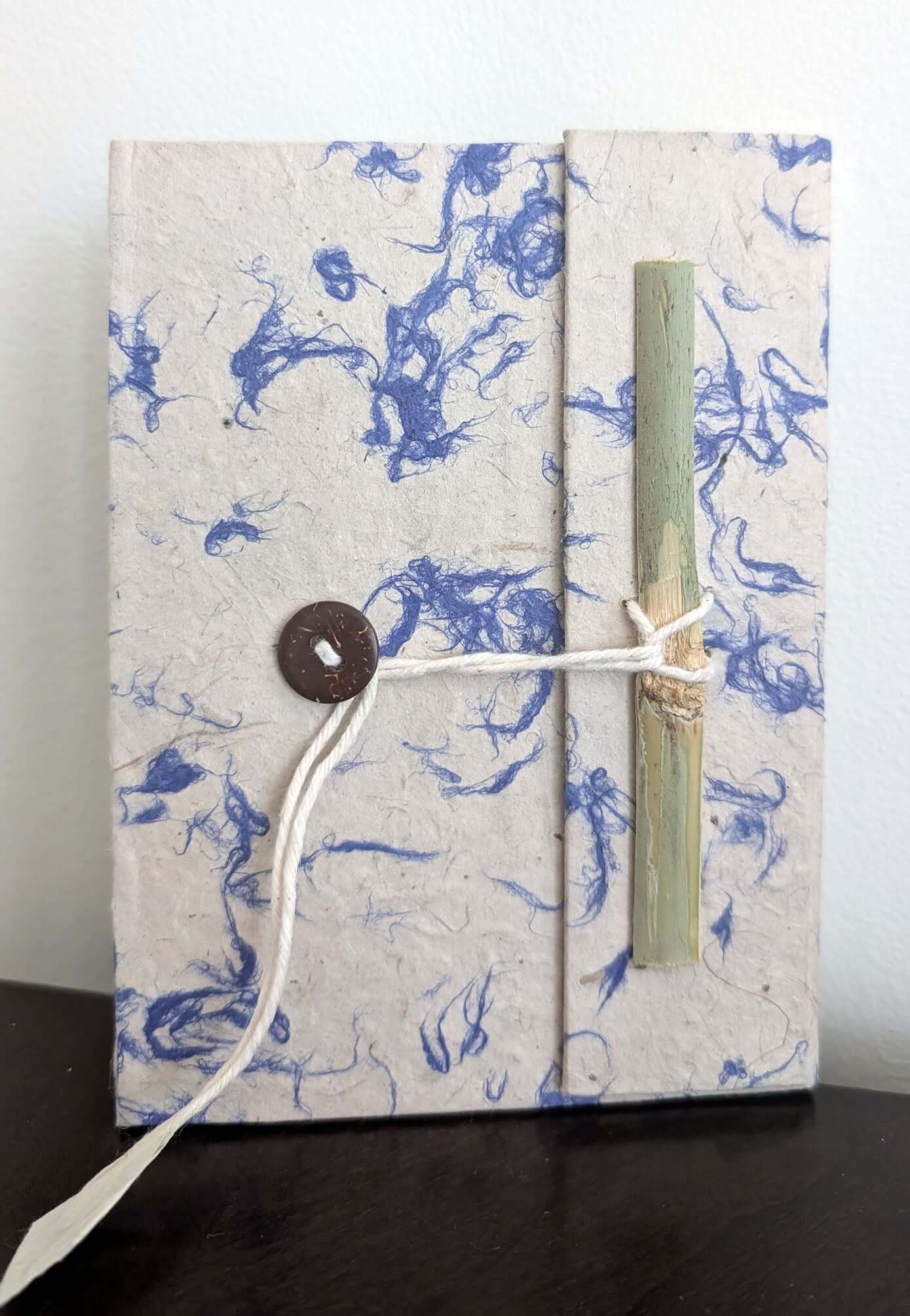 A handmade Lokta paper notebook with blue fiber detailing, secured with a string-and-bamboo tie, standing upright on a dark surface.
