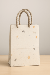 A handcrafted Lokta paper gift bag with twisted natural handles, displayed standing on a dark wooden surface. The bag features a textured, off-white base with scattered botanical inclusions in orange, lavender, and green tones, showcasing its natural, eco-friendly aesthetic.