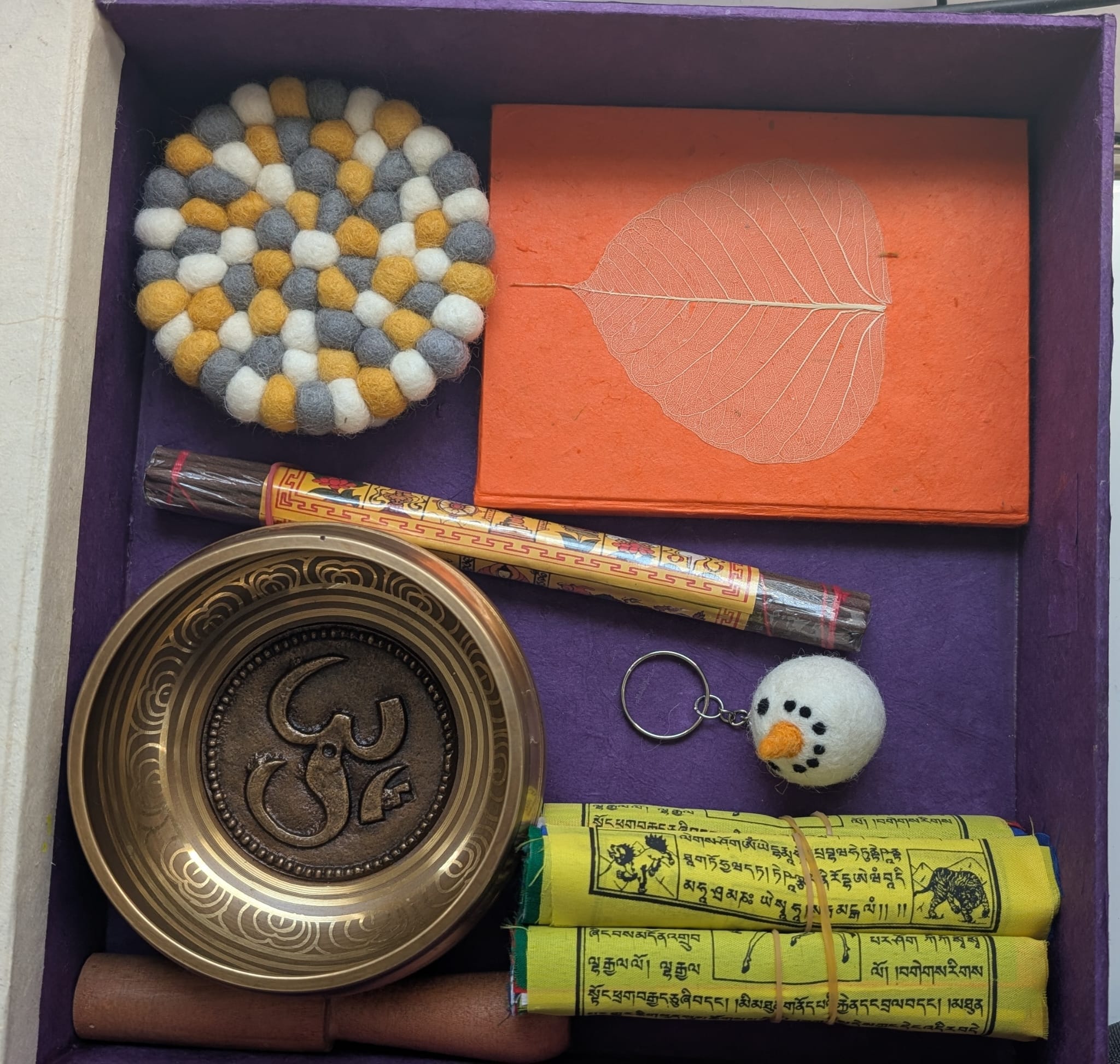 A warm lifestyle flat lay featuring a purple MAHA Treasures gift box surrounded by Nepali handmade items, including a brass singing bowl with an elephant design, an orange Lokta journal with a Bodhi leaf, rolled prayer flags, incense sticks, a felt snowman keychain, and a felt ball coaster.