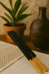 A neat bundle of black incense sticks with golden bamboo bases arranged on a soft beige mat, with a terracotta pot and earthenware vase in the blurred background.