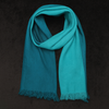 Two-tone teal merino wool stole with soft gradient shading and fringed edges, draped in a loop against a dark textured background – handwoven in Nepal.