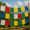 Five strands of colorful Tibetan prayer flags—each with white, red, green, yellow, and blue cotton flags—hang between bamboo poles outdoors, fluttering against a clear sky and green mountain landscape. Each flag is printed with sacred Tibetan mantras and spiritual symbols.