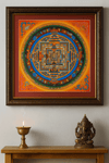 A framed Tibetan Buddhist mandala thangka painting with intricate geometric patterns in vibrant orange, blue, green, and gold tones hangs on a light-colored wall. Below it, a wooden shelf holds a lit brass butter lamp emitting a thin trail of smoke and a small brass statue of the Hindu deity Ganesha.