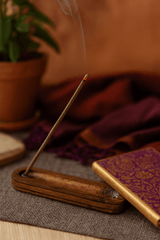A smoldering bundle of Tibetan rope incense rests on a wooden tray, with soft smoke rising and a blurred potted plant in the background.